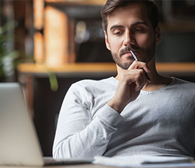 White middle age man with beard and wearing a grey long sleeve shirt holds a pencil to his mouth pensively while looking at an open laptop on a desk in front of him.