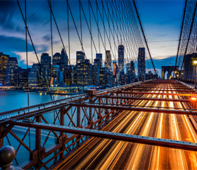 Long-exposure photograph of a suspension bridge leading to a city lit by a sunset and city lights.