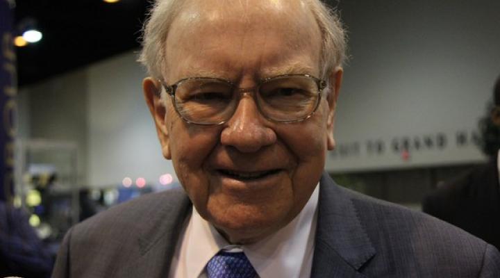 Warren Buffett wearing a gray suit, blue tie, and smiling into the camera