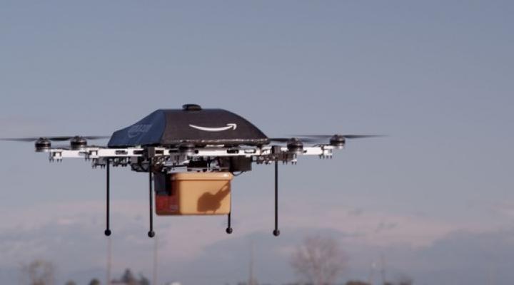 An Amazon-branded drone mid-flight holding a package in a yellow container. 