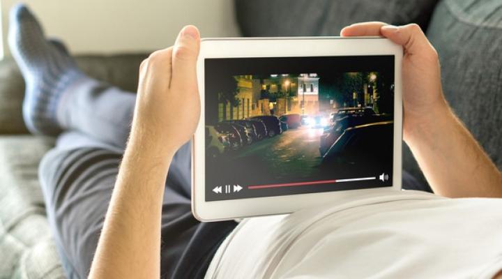 A man laying on a couch watching streaming video on a tablet. 