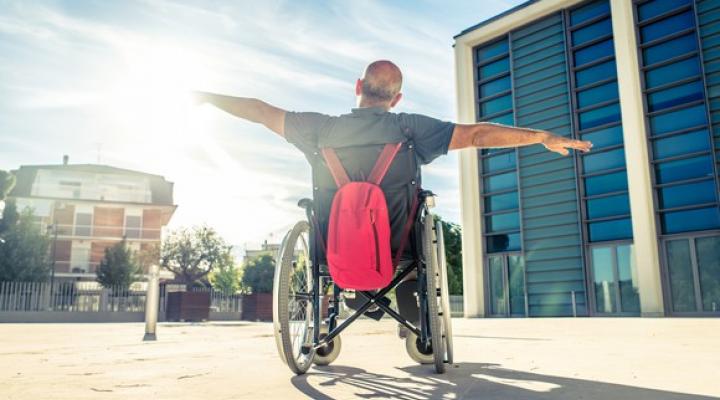 A man in a wheelchair outside of a building raises his arms vertically as if to fly.