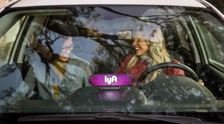 Two smiling women sitting in the front seat of a car with the Lyft logo mounted on the dashboard