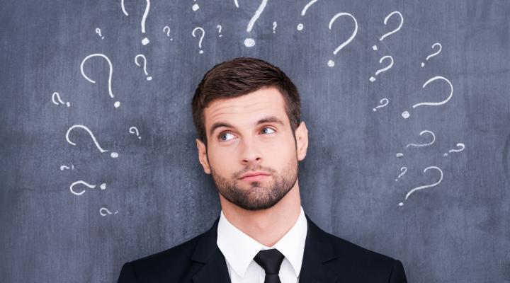 a young man in a suit stands in front of a blackboard with question marks written above his head.