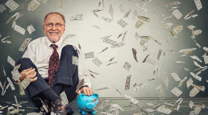 An older businessman sits against a wall with money falling down around him.