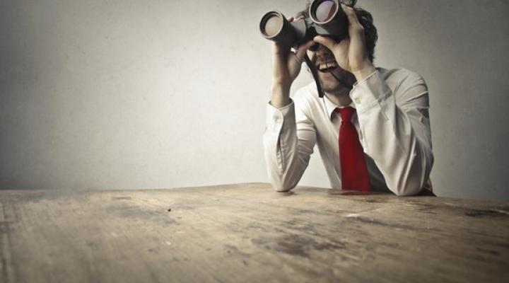 A businessman sitting at a large table and looking through binoculars.