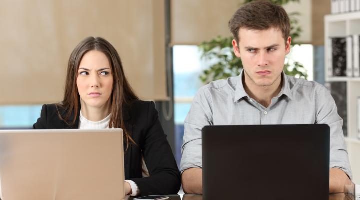 A woman and a man shoot each other looks as they sit side by side at laptops.