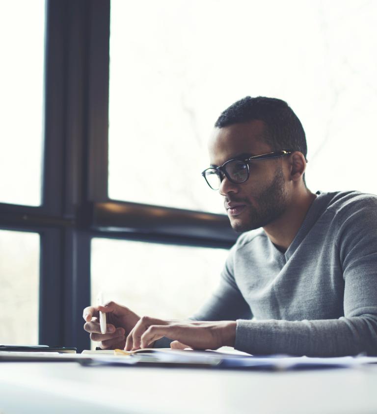 A man working on a notebook computer at a table with large windows next to him.